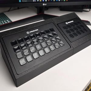 Peut inclure: Une console de contrôle Stream Deck noire dotée d'une grande grille de trente-deux boutons carrés sur la gauche et d'une plus petite grille de quinze boutons carrés sur la droite, les deux sections portant le logo Stream Deck en blanc.