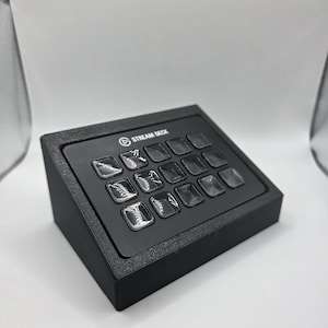 May include: A black Stream Deck control console featuring a slanted design with twelve square, translucent buttons arranged in a grid. The device has a matte black finish and the text STREAM DECK is printed in white on the top center panel.