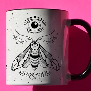 May include: White ceramic mug with a black handle and rim. The mug features a black and white illustration of an eye and a moth with intricate details. The mug is set against a bright pink background.