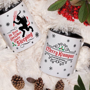 May include: Two white ceramic mugs with black rims. One mug features a black silhouette of Krampus with the text "He Sees You When You're Sleeping". The other mug has the text "Merry Krampus" with the words "Gruss Uom Krampus" underneath.