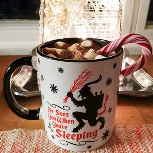 May include: White ceramic mug with a black handle and a black silhouette of a creature with horns and a tail. The mug has red and green holly and snowflakes printed on it. The text on the mug reads "He Sees You When You're Sleeping". The mug is filled with hot chocolate and marshmallows and a candy cane is sticking out of the mug.