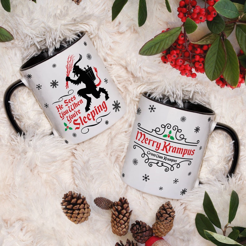 May include: Two white ceramic mugs with black rims. One mug features a black silhouette of Krampus with the text "He Sees You When You're Sleeping". The other mug has the text "Merry Krampus" with the words "Gruss Uom Krampus" underneath.