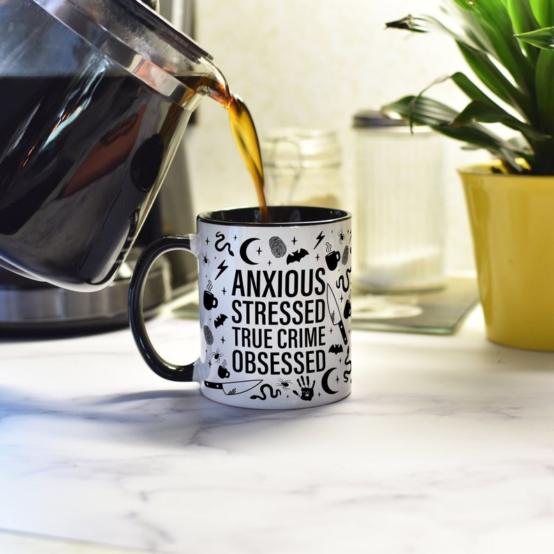 May include: A white coffee mug with a black rim and handle. The mug has black text that reads "Anxious Stressed True Crime Obsessed" and is surrounded by various black icons, including bats, spiders, snakes, and crescent moons.