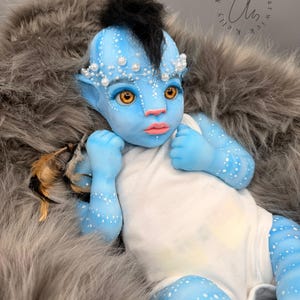 Może przedstawiać: Niebieskoskóra lalka dla dziecka z białymi kropkami, dużymi żółtymi oczami i czarnym irokezem. Lalka ma na sobie białe body i opaskę na głowę ozdobioną perłami. Spoczywa na szarym sztucznym futrze. Widoczny jest podpis artysty.