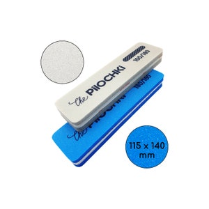 Puede incluir: Dos limas de uñas, una blanca y otra azul, apiladas sobre un fondo blanco. La lima blanca tiene el texto "The Piiochki" y "100/180" impreso. La lima azul tiene el texto "180/180" y "The Piiochki" impreso. Una incrustación circular muestra la textura de la lima. Las dimensiones son 115 x 140 mm.