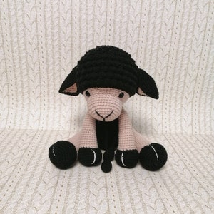 May include: A crocheted black and pink sheep plush toy with black eyes and a black nose. The sheep is sitting with its legs crossed and its tail curled up.