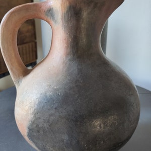 Op de afbeelding: Een handgemaakte, rustieke kan met een afgeronde body en een gebogen handvat. De kan is voornamelijk donkergrijs met delen roodbruin en zwart. Hij heeft een tuit om uit te schenken. De kan is waarschijnlijk van klei.