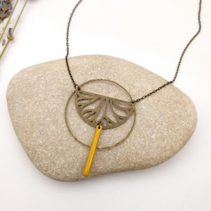 May include: A gold necklace with a geometric pendant featuring a circle and a half-circle with a detailed design. The pendant is accented with a yellow stick.