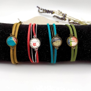 Könnte beinhalten: Fünf Armbänder mit verschiedenen farbigen Schnüren und runden Glasanhängern. Die Anhänger haben unterschiedliche Muster und Farben, darunter Blau, Rot, Grün und Gold.