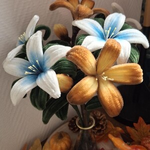 Op de afbeelding: Een boeket kunstbloemen in een kleine vaas. De bloemen zijn wit met blauwe accenten en oranjebruin. Groene bladeren omringen de bloemen. De opstelling staat op een houten basis met herfstdecoraties, waaronder bladeren en kalebassen.