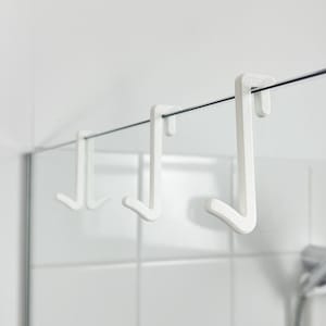 Op de afbeelding: Drie witte plastic haken hangen aan een douchestang in een badkamer. De haken zijn ontworpen om spullen over de douchedeur te hangen.