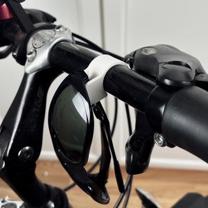 Può includere: Un manubrio di bicicletta nero con un portaocchiali in plastica bianca, un supporto per telefono nero e leve dei freni nere.
