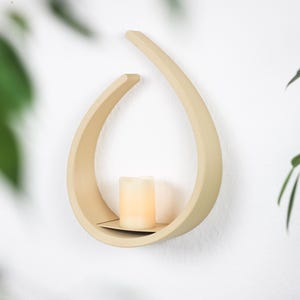 Può includere: Un ripiano a muro in legno beige a forma di lacrima con un piccolo ripiano per esporre una candela. La candela è di colore crema e si trova sul ripiano.