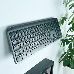 Könnte beinhalten: Eine schwarze drahtlose Tastatur mit runden Tasten ist an einer weißen Wand montiert. Die Tastatur ist leicht nach unten geneigt.