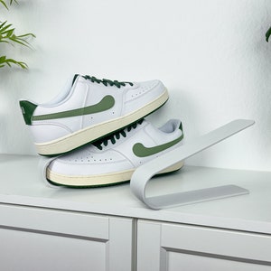 Peut inclure: Deux baskets blanches avec des accents verts sont présentées sur un support à chaussures blanc. Les baskets ont des lacets blancs et une semelle blanche.