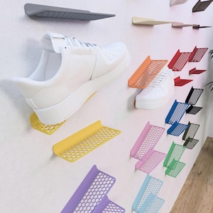 Può includere: Un espositore da parete con mensole geometriche in metallo colorate con un motivo a nido d'ape. Le mensole sono progettate per contenere scarpe e altri piccoli oggetti. Una scarpa bianca è esposta su una delle mensole.