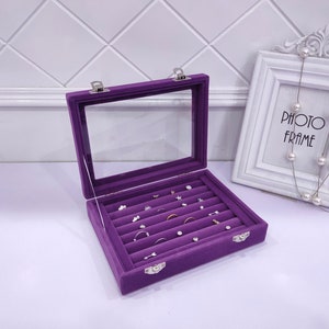 May include: Purple velvet jewelry box with a clear lid and multiple compartments for rings and earrings. The box is open and the compartments are filled with jewelry.