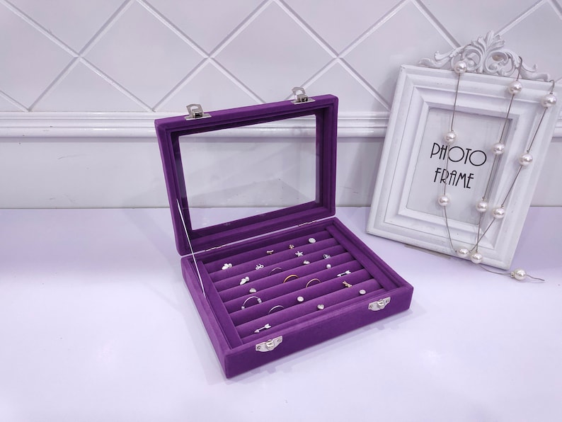 May include: Purple velvet jewelry box with a clear lid and multiple compartments for rings and earrings. The box is open and the compartments are filled with jewelry.