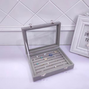 May include: A gray velvet jewelry box with a clear lid and multiple compartments for storing rings and earrings. The box has two silver clasps.