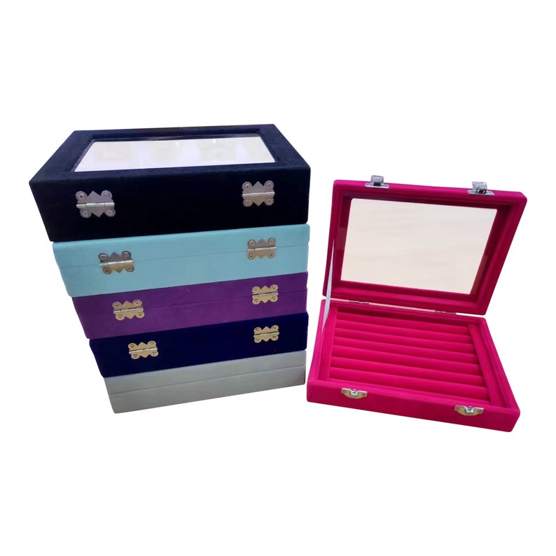 May include: Stack of five velvet ring boxes with clear lids. The top box is black, the second is light blue, the third is purple, the fourth is dark blue, and the bottom box is light blue. The boxes have gold hinges and are lined with velvet.