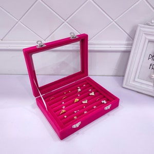 May include: A pink velvet jewelry box with a clear lid and multiple compartments for rings and earrings. The box has silver clasps and is filled with various gold and silver jewelry.