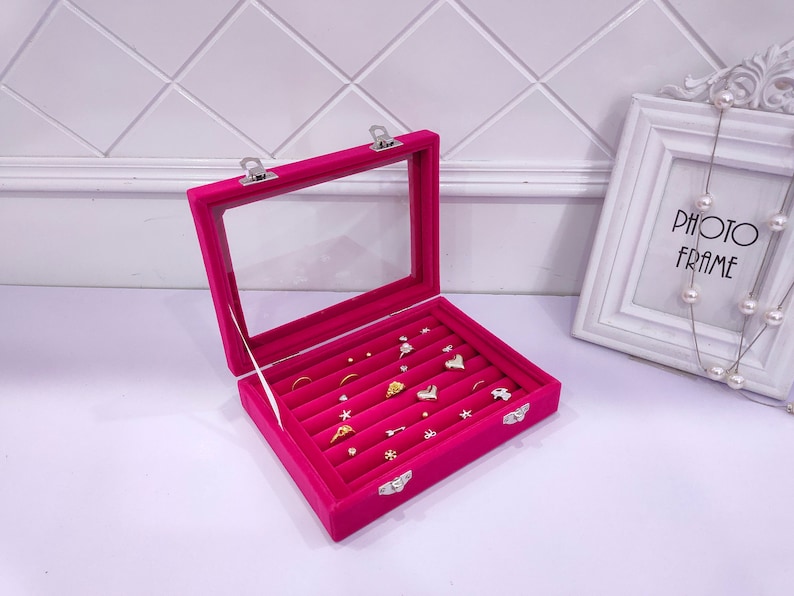 May include: A pink velvet jewelry box with a clear lid and multiple compartments for rings and earrings. The box has silver clasps and is filled with various gold and silver jewelry.