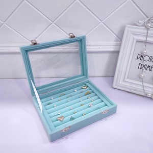 May include: A light blue velvet jewelry box with a clear lid. The box has multiple rows of compartments for storing rings and earrings. The box is open and there are several pieces of jewelry inside.