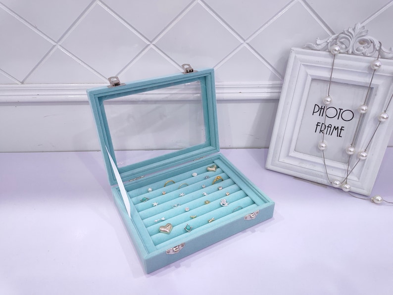 May include: A light blue velvet jewelry box with a clear lid. The box has multiple rows of compartments for storing rings and earrings. The box is open and there are several pieces of jewelry inside.