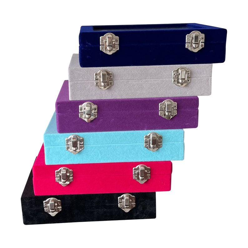 May include: Six velvet jewelry boxes stacked on top of each other. The boxes are in different colors: black, red, light blue, purple, gray, and navy blue. Each box has a silver latch.