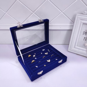 May include: A blue velvet ring box with a clear lid and multiple compartments for storing rings and other jewelry. The box is open and has several rings inside.