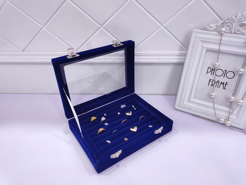 May include: A blue velvet ring box with a clear lid and multiple compartments for storing rings and other jewelry. The box is open and has several rings inside.
