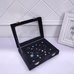 May include: Black velvet jewelry box with a clear lid and multiple compartments for storing earrings, rings, and other small jewelry. The box is closed and sitting on a white surface.