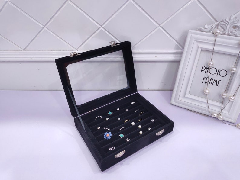 May include: Black velvet jewelry box with a clear lid and multiple compartments for storing earrings, rings, and other small jewelry. The box is closed and sitting on a white surface.