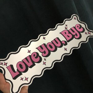 以下が含まれることがあります： 波状の縁取りのある長方形のサインで、テキスト「Love you, Bye」がピンク色で黒で縁取られています。小さな星のグラフィックがテキストの周りに散らばっています。サインは白い背景で、濃い緑色の背景に対して保持されています。