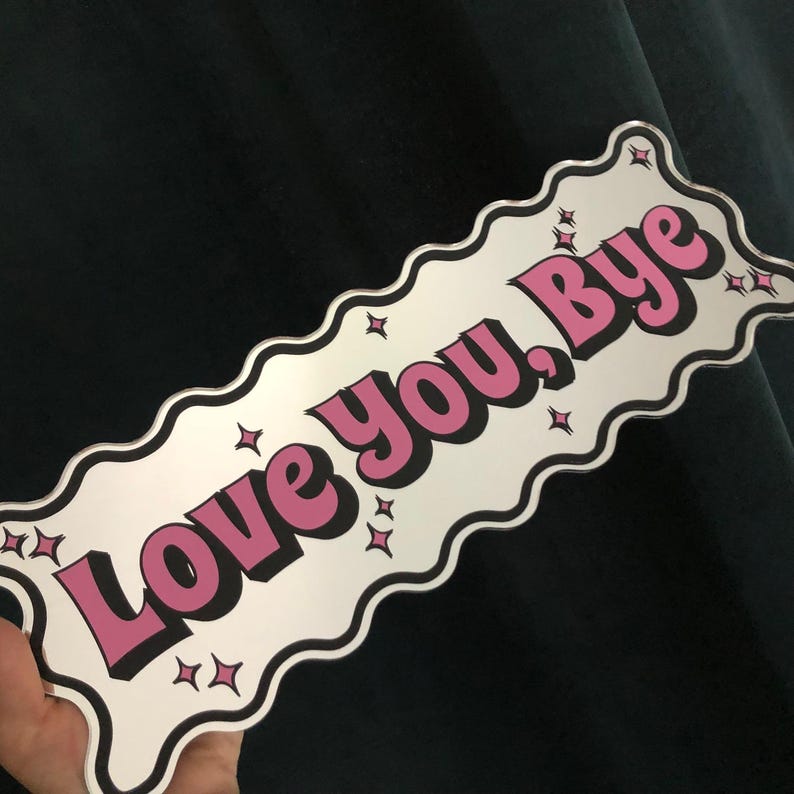 以下が含まれることがあります： 波状の縁取りのある長方形のサインで、テキスト「Love you, Bye」がピンク色で黒で縁取られています。小さな星のグラフィックがテキストの周りに散らばっています。サインは白い背景で、濃い緑色の背景に対して保持されています。