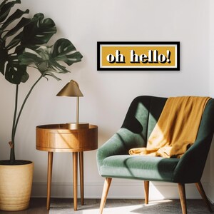 May include: A black framed print with a yellow background and the text "oh hello!" in white letters. The print is hanging on a white wall above a green armchair with a yellow throw blanket.