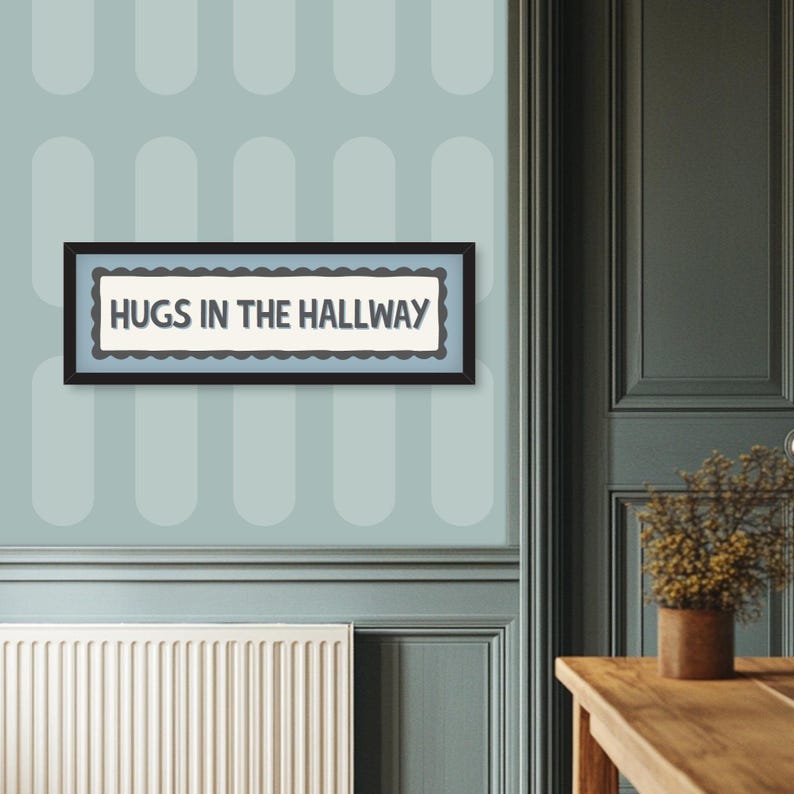 May include: A framed sign with the text "HUGS IN THE HALLWAY" in a black frame. The sign has a light blue background with a scalloped border. The wall behind the sign is light blue with a repeating pattern.