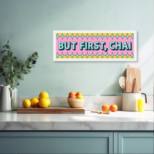 May include: A white framed print with a pink background and yellow and blue geometric shapes. The text "BUT FIRST, CHAI" is written in blue and white letters.