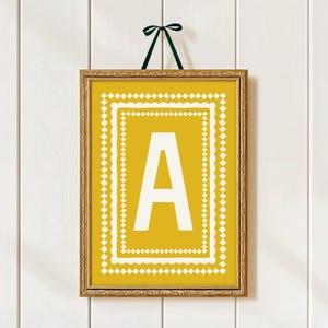 May include: A framed print with the letter "A" in white on a yellow backdrop. The print has a white diamond-patterned border. The frame is gold-coloured, and a green ribbon is used for hanging. The print is suitable for home decor.