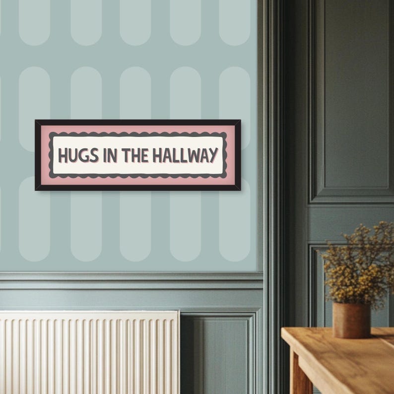 May include: A framed rectangular sign with the text "HUGS IN THE HALLWAY" in a black frame. The sign has a pink border with a scalloped edge and is mounted on a light blue wall with a repeating oval pattern.