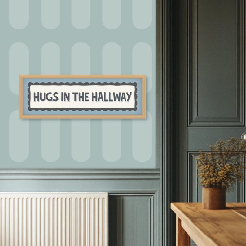 May include: A framed sign with the text "HUGS IN THE HALLWAY" in a light blue and white color scheme. The sign has a scalloped border and is set against a light blue wall with a repeating oval pattern. The frame is a light brown color.