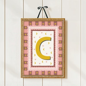 May include: A framed art print with the letter "C" in mustard yellow. The artwork has a pink and brown patterned background with stars and crescent moons. The gold frame is suspended by a green ribbon.