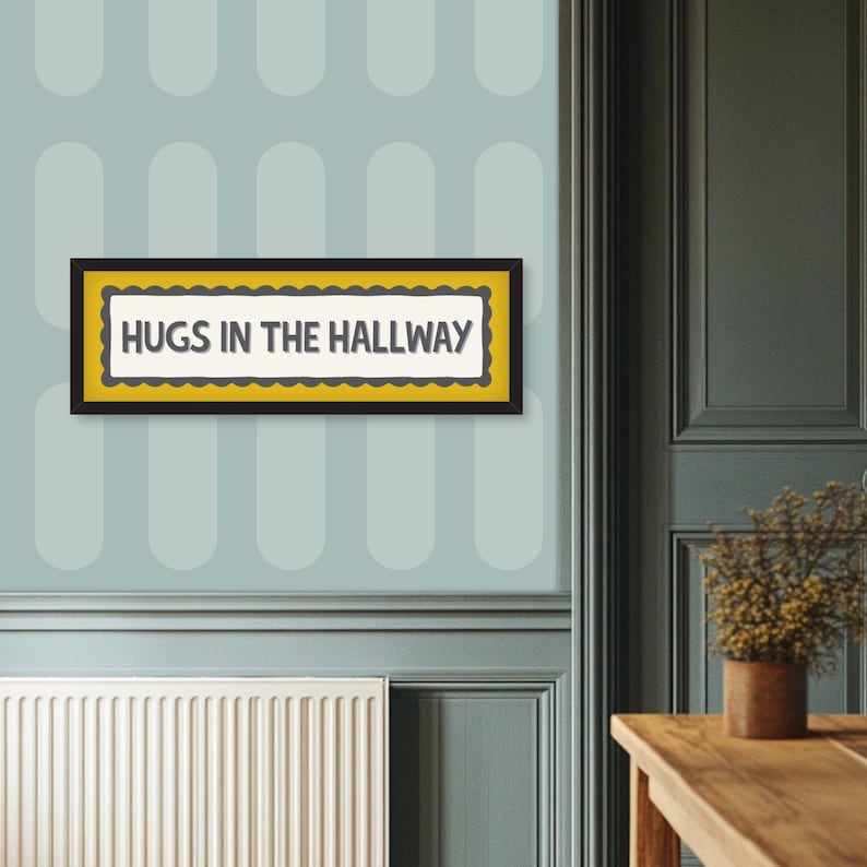 May include: A rectangular wall art piece with the text "HUGS IN THE HALLWAY" in a black frame. The artwork has a yellow border with a scalloped edge and is set against a light blue wall with a repeating oval pattern.