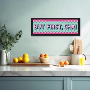 May include: A black framed sign with a pink and blue background and the text "BUT FIRST, CHAI" in pink letters. The sign is hanging on a light blue wall in a kitchen setting.