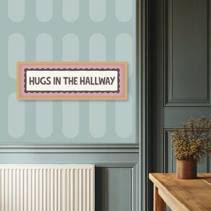 May include: A framed sign with the text "HUGS IN THE HALLWAY" in a light pink and brown frame. The sign is mounted on a light blue wall with a repeating oval pattern. The wall is next to a dark green door.