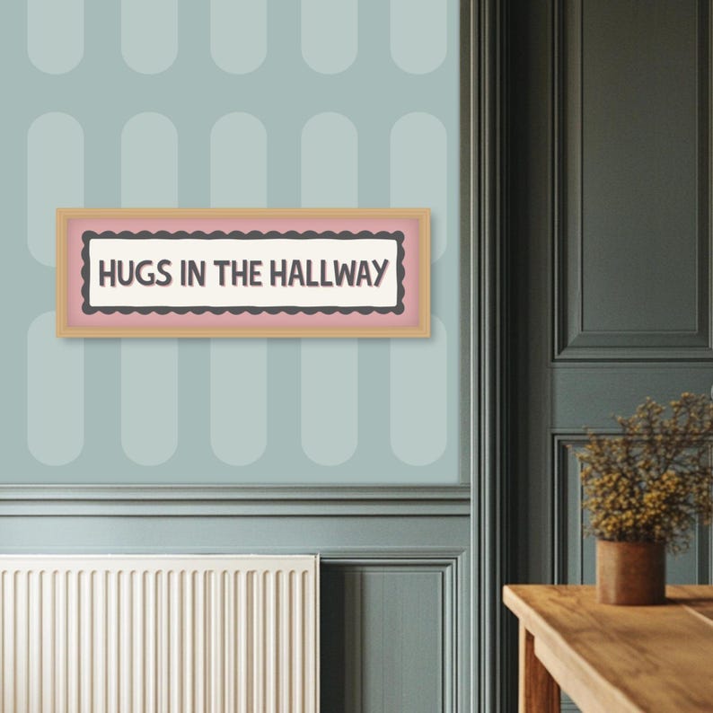 May include: A framed sign with the text "HUGS IN THE HALLWAY" in a light pink and brown frame. The sign is mounted on a light blue wall with a repeating oval pattern. The wall is next to a dark green door.