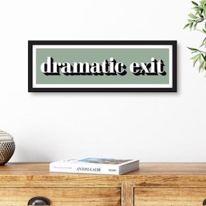 May include: A framed print featuring the words "dramatic exit" in a bold, white font with a black shadow effect. The text is set against a sage green background, framed in black. The print is displayed on a wooden surface.