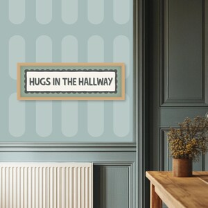 May include: A framed sign with the text "HUGS IN THE HALLWAY" in a black font. The sign has a light brown frame and a scalloped border. The sign is hanging on a light blue wall with a repeating oval pattern.