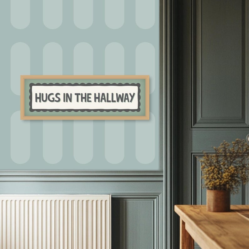 May include: A framed sign with the text "HUGS IN THE HALLWAY" in a black font. The sign has a light brown frame and a scalloped border. The sign is hanging on a light blue wall with a repeating oval pattern.