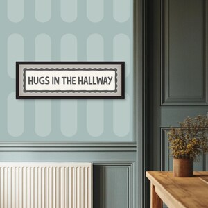 May include: A rectangular framed sign with the text "HUGS IN THE HALLWAY" in black letters. The sign has a scalloped border and is set against a light background. The frame is black. The sign is hanging on a light blue wall.